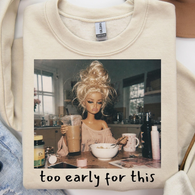 Peut inclure: Un sweat-shirt beige &agrave; col rond avec une impression graphique repr&eacute;sentant une poup&eacute;e aux cheveux blonds en chignon d&eacute;coiff&eacute;, assise &agrave; une table avec un caf&eacute; et un bol de c&eacute;r&eacute;ales. Le texte sous l'image indique "too early for this" en noir.