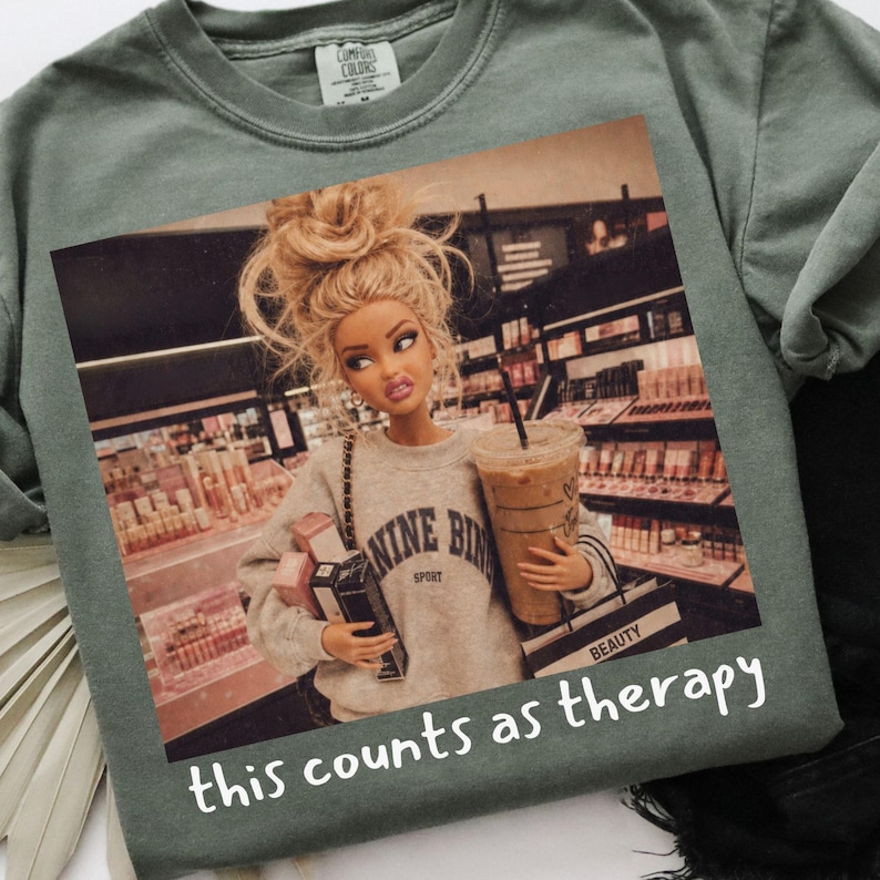 Op de afbeelding: Een saliegroen t-shirt met een grafische print van een blonde pop met een knotje, gekleed in een grijs sweatshirt met de tekst NINE BINE SPORT. De pop houdt een koffiebeker en winkeltassen vast in een winkel. De tekst this counts as therapy staat eronder.