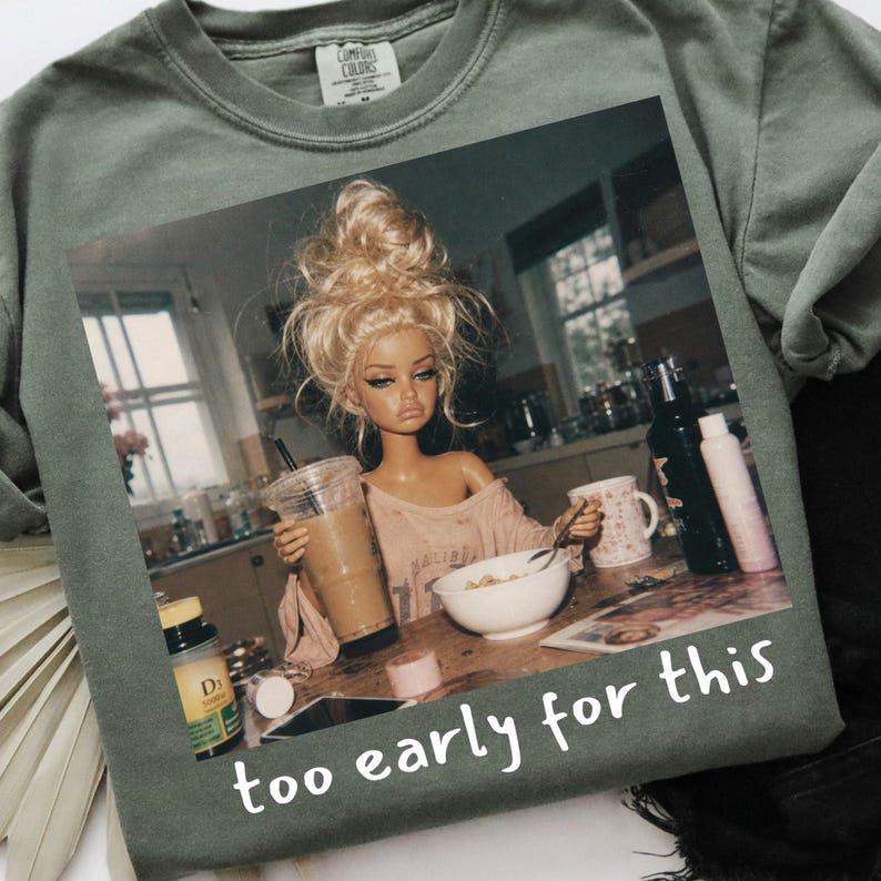 Peut inclure: Un t-shirt vert sauge avec une impression graphique repr&eacute;sentant une poup&eacute;e aux cheveux blonds en chignon d&eacute;coiff&eacute;, assise &agrave; une table de cuisine avec un caf&eacute; et un bol de c&eacute;r&eacute;ales. Le texte "too early for this" est imprim&eacute; en blanc en bas.