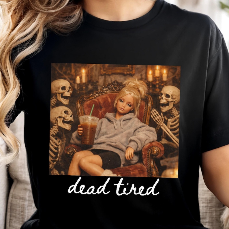 K&ouml;nnte beinhalten: Ein schwarzes T-Shirt mit einem Grafikdruck einer blonden Puppe im grauen Kapuzenpullover, die in einem Sessel sitzt und einen Eiskaffee h&auml;lt, flankiert von zwei Skeletten. Darunter steht der Text "dead tired" in wei&szlig;er Schreibschrift.