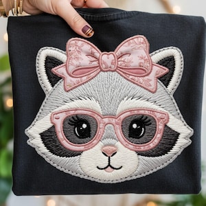 Könnte beinhalten: Schwarzes Sweatshirt mit einem gestickten Waschbär-Gesicht. Der Waschbär hat graues und weißes Fell, eine rosa Brille und eine rosa Schleife mit Blumenmuster. Das Design befindet sich mittig auf der Vorderseite des Sweatshirts.