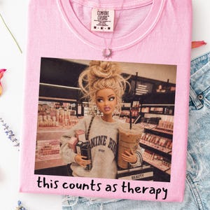 Op de afbeelding: Een lichtroze t-shirt met een grafische print van een blonde pop met een rommelige knot in een grijs sweatshirt, die boodschappentassen en een ijskoffie vasthoudt in een winkel. De tekst "this counts as therapy" staat onder de afbeelding.