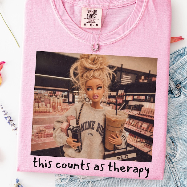 Op de afbeelding: Een lichtroze t-shirt met een grafische print van een blonde pop met een rommelige knot in een grijs sweatshirt, die boodschappentassen en een ijskoffie vasthoudt in een winkel. De tekst "this counts as therapy" staat onder de afbeelding.
