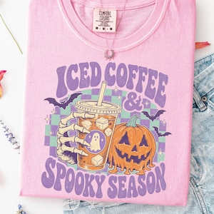 May include: A light pink t-shirt featuring a graphic print of a skeleton hand holding an iced coffee cup next to a melting jack-o'-lantern. The design includes purple text reading ICED COFFEE & SPOOKY SEASON, a green and purple checkerboard, and bats.
