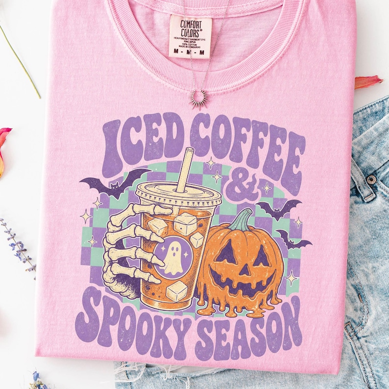 May include: A light pink t-shirt featuring a graphic print of a skeleton hand holding an iced coffee cup next to a melting jack-o'-lantern. The design includes purple text reading ICED COFFEE & SPOOKY SEASON, a green and purple checkerboard, and bats.