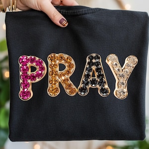 Puede incluir: Sudadera negra con la palabra "PRAY" escrita en letras grandes y enjoyadas. Las letras están delineadas en dorado y rellenas de cristales rosas, ámbar, negros y transparentes. La sudadera es sostenida por una mano.