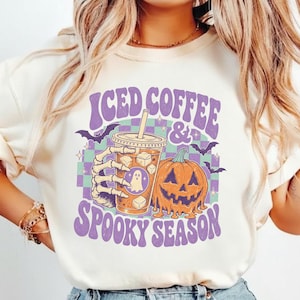 May include: A cream-colored t-shirt featuring a graphic of a skeleton hand holding an iced coffee cup next to a jack-o'-lantern. The design includes purple text that reads ICED COFFEE & SPOOKY SEASON, small bats, and a green and purple checkerboard background.
