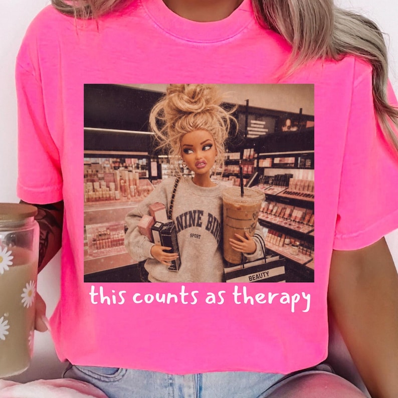Op de afbeelding: Een neonroze t-shirt met een grafische print van een pop met blond haar in een knot, gekleed in een grijze sweater, met winkeltassen en een ijskoffie in een winkel. De tekst onder de afbeelding luidt, this counts as therapy.
