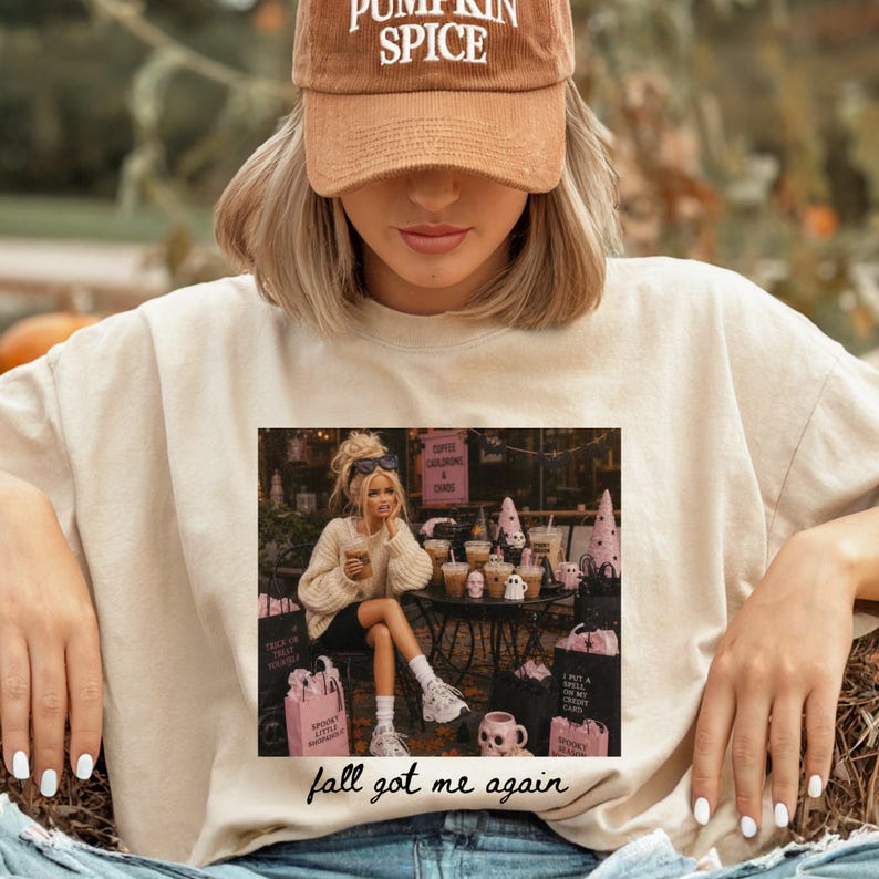 May include: A beige crewneck sweatshirt featuring a graphic print of a person sitting at a table with pink Halloween-themed decor and drinks. Below the graphic, the text reads "fall got me again" in black cursive script. A brown corduroy baseball cap with "PUMPKIN SPICE" embroidered in white text is also visible.
