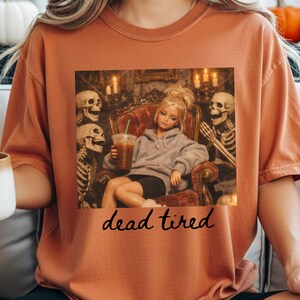 K&ouml;nnte beinhalten: Ein rostorangefarbenes T-Shirt mit einem Grafikdruck einer blonden Puppe in einem grauen Kapuzenpullover und schwarzen Shorts, die in einem Sessel sitzt, einen Eiskaffee h&auml;lt und von zwei Skeletten flankiert wird. Darunter steht "dead tired".