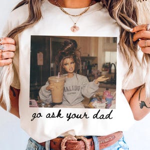 Op de afbeelding: Cr&egrave;mekleurig T-shirt met een afbeelding van een pop die een koffiedrank vasthoudt, met een grijze sweatshirt met de tekst "Malibu California". De tekst "go ask your dad" is eronder gedrukt. Een casual kledingstuk voor dagelijks gebruik.