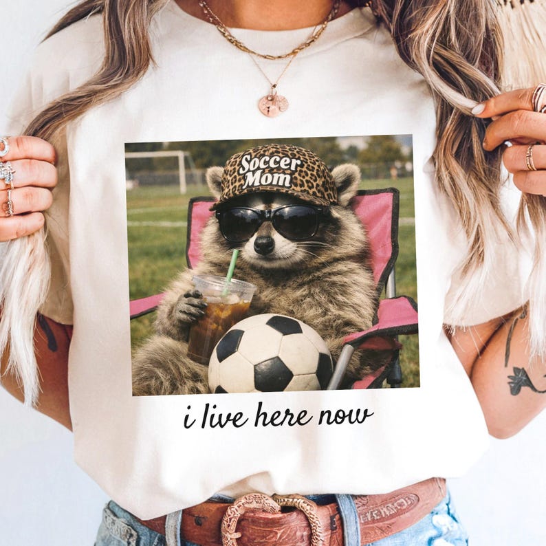Puede incluir: Camiseta blanca con un mapache con gafas de sol y un sombrero con estampado de leopardo que dice "Soccer Mom". El mapache sostiene una bebida y est&aacute; sentado en una silla rosa junto a un bal&oacute;n de f&uacute;tbol. El texto "i live here now" est&aacute; debajo.