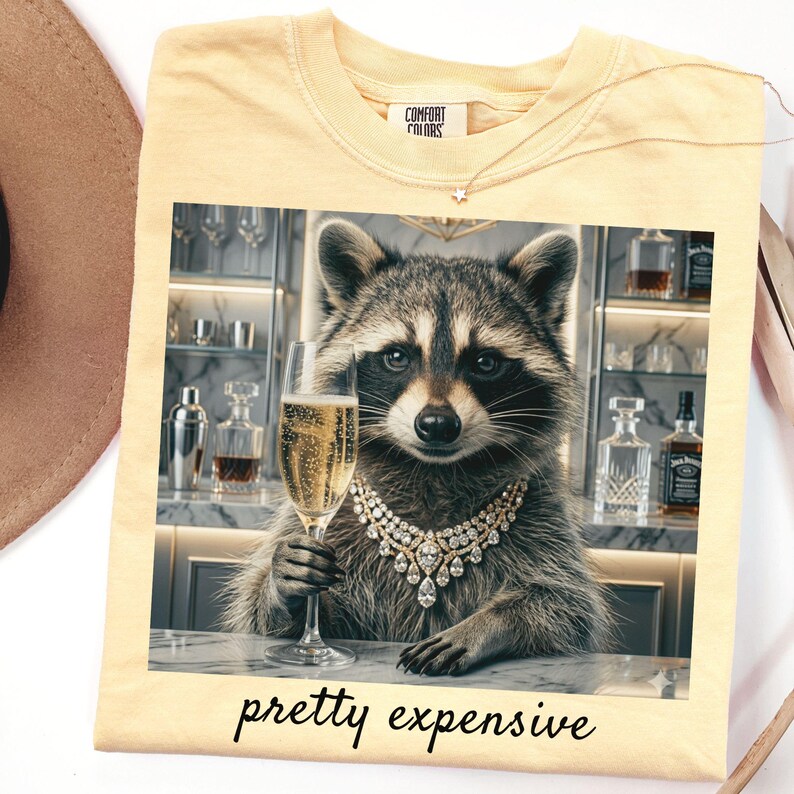May include: Yellow t-shirt with a raccoon graphic wearing a diamond necklace and holding a champagne glass. The image has the text "pretty expensive" at the bottom. The shirt has a Comfort Colors tag.