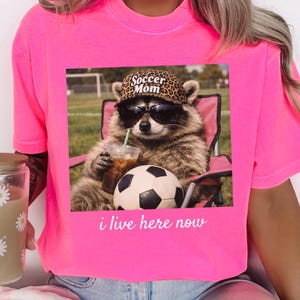 Puede incluir: Camiseta rosa brillante con un gr&aacute;fico de un mapache con gafas de sol y un sombrero con estampado de leopardo que dice "Soccer Mom". El mapache est&aacute; sentado en una silla rosa, sosteniendo una bebida y un bal&oacute;n de f&uacute;tbol. El texto "i live here now" est&aacute; debajo.