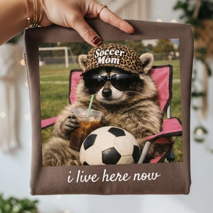 Puede incluir: Un art&iacute;culo decorativo cuadrado marr&oacute;n con un mapache con gafas de sol y un sombrero con estampado de leopardo que dice "Soccer Mom". El mapache est&aacute; sentado en una silla rosa, sosteniendo una bebida y junto a un bal&oacute;n de f&uacute;tbol. El texto "i live here now" est&aacute; en la parte inferior.