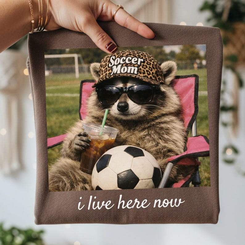 Puede incluir: Un art&iacute;culo decorativo cuadrado marr&oacute;n con un mapache con gafas de sol y un sombrero con estampado de leopardo que dice "Soccer Mom". El mapache est&aacute; sentado en una silla rosa, sosteniendo una bebida y junto a un bal&oacute;n de f&uacute;tbol. El texto "i live here now" est&aacute; en la parte inferior.