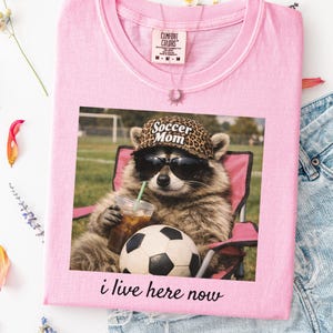 Puede incluir: Camiseta rosa claro con un estampado de un mapache con gafas de sol y un sombrero con estampado de leopardo, sosteniendo una bebida y sentado en una silla rosa junto a un bal&oacute;n de f&uacute;tbol. El texto "Soccer Mom" est&aacute; en el sombrero, y "i live here now" est&aacute; debajo.