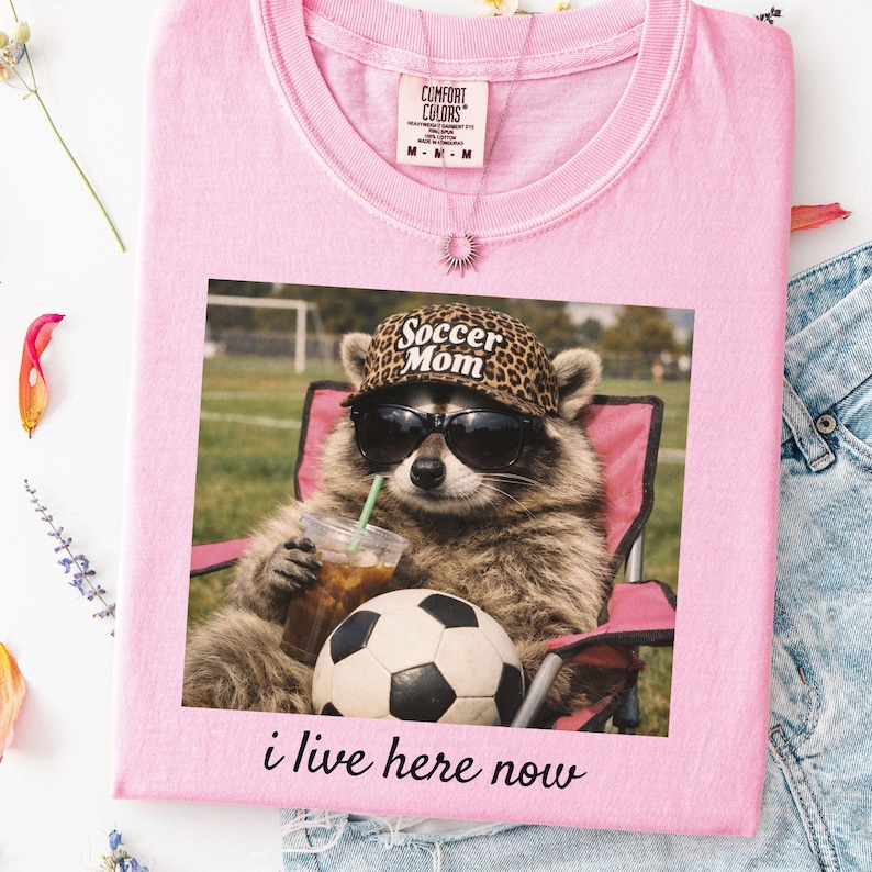 Puede incluir: Camiseta rosa claro con un estampado de un mapache con gafas de sol y un sombrero con estampado de leopardo, sosteniendo una bebida y sentado en una silla rosa junto a un bal&oacute;n de f&uacute;tbol. El texto "Soccer Mom" est&aacute; en el sombrero, y "i live here now" est&aacute; debajo.
