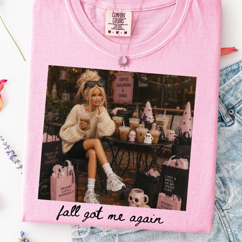 May include: A light pink t-shirt featuring a graphic print of a doll with blonde hair wearing a cream sweater and black shorts, sitting at a table with coffee cups and Halloween decor. The text "fall got me again" is printed in black below the graphic.