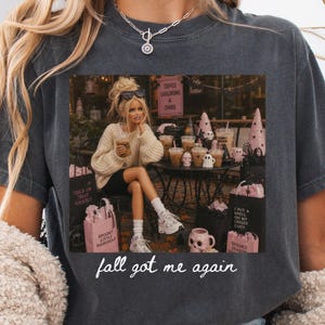 May include: A charcoal gray t-shirt featuring a graphic of a blonde doll in a cream sweater sitting at a table with coffee cups, pink shopping bags, and skull decor. The text reads "COFFEE CAULDRONS & CHAOS", "TRICK OR TREAT YOURSELF", "I PUT A SPELL ON MY CREDIT CARD", "SPOOKY LITTLE SHOPAHOLIC", "SPOOKY SEASON", and "fall got me again".