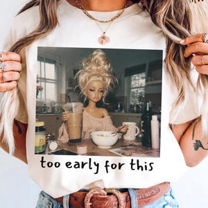 Peut inclure: Un t-shirt couleur cr&egrave;me avec une impression graphique repr&eacute;sentant une poup&eacute;e blonde en chignon d&eacute;sordonn&eacute; assise &agrave; une table de cuisine avec un caf&eacute; et un bol de c&eacute;r&eacute;ales. Le texte "too early for this" est imprim&eacute; en noir sous l'image.
