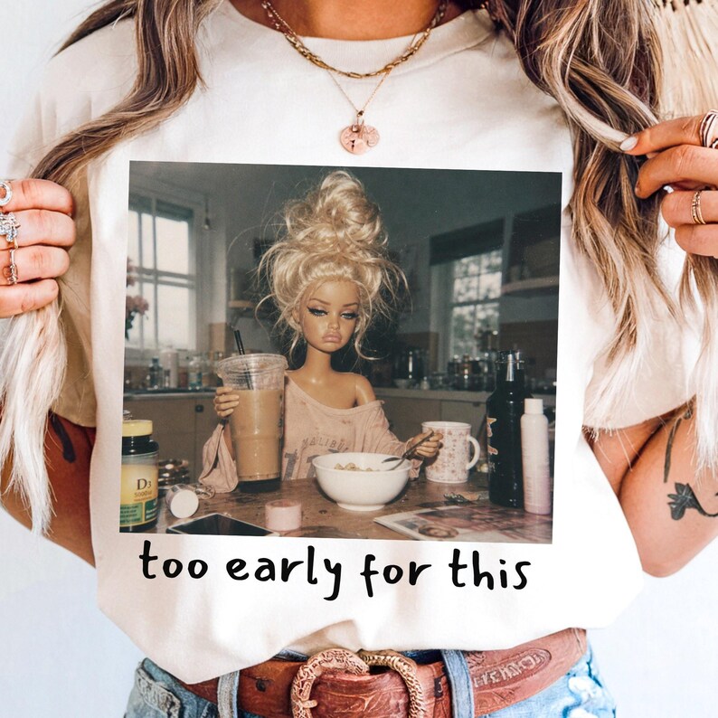 Peut inclure: Un t-shirt couleur cr&egrave;me avec une impression graphique repr&eacute;sentant une poup&eacute;e blonde en chignon d&eacute;sordonn&eacute; assise &agrave; une table de cuisine avec un caf&eacute; et un bol de c&eacute;r&eacute;ales. Le texte "too early for this" est imprim&eacute; en noir sous l'image.