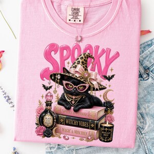 May include: A light pink t-shirt featuring a graphic of a black cat wearing a leopard print witch hat and pink glasses. The cat rests on three books labeled Spells & Potions, Witchy Vibes, and Magic & Mischief, surrounded by roses, a potion bottle, and a candle.