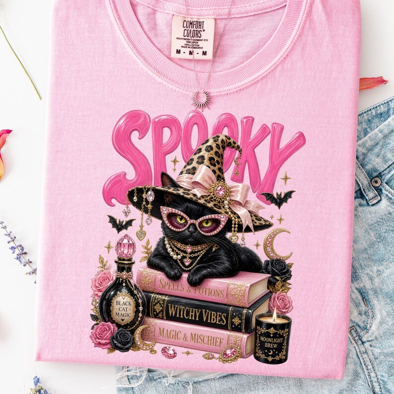 May include: A light pink t-shirt featuring a graphic of a black cat wearing a leopard print witch hat and pink glasses. The cat rests on three books labeled Spells & Potions, Witchy Vibes, and Magic & Mischief, surrounded by roses, a potion bottle, and a candle.