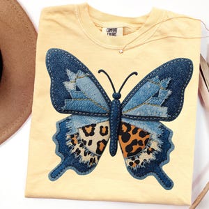 May include: A light yellow Comfort Colors t-shirt featuring a large butterfly design. The butterfly has denim blue wings with a leopard print pattern on the lower wings. A small gold necklace with a star pendant is visible.