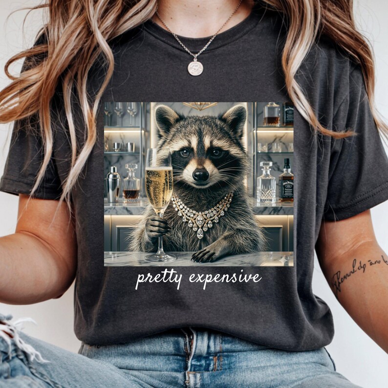 May include: Dark gray t-shirt featuring a raccoon wearing a diamond necklace and holding a champagne glass. The raccoon is behind a bar with bottles and the text "pretty expensive".