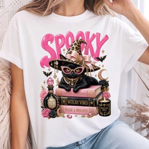 May include: A white t-shirt featuring a graphic of a black cat wearing a leopard print witch hat, pink glasses, and jewelry. The cat rests on three books labeled Spells & Potions, Witchy Vibes, and Magic & Mischief, with pink roses and text.