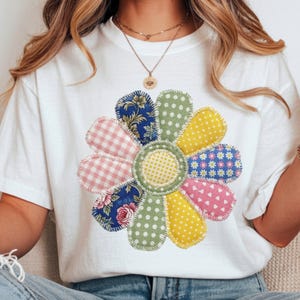 May include: White t-shirt featuring a large, colorful flower design. The flower is made of various fabric patterns, including gingham, polka dots, and floral prints in shades of pink, blue, yellow, and green. The design is centered on the shirt.