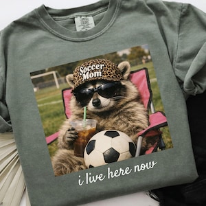 Puede incluir: Camiseta Comfort Colors verde salvia con un gr&aacute;fico de un mapache con gafas de sol y un sombrero de leopardo que dice "Soccer Mom". El mapache est&aacute; sentado en una silla rosa, sosteniendo una bebida y un bal&oacute;n de f&uacute;tbol. El texto "i live here now" est&aacute; debajo.
