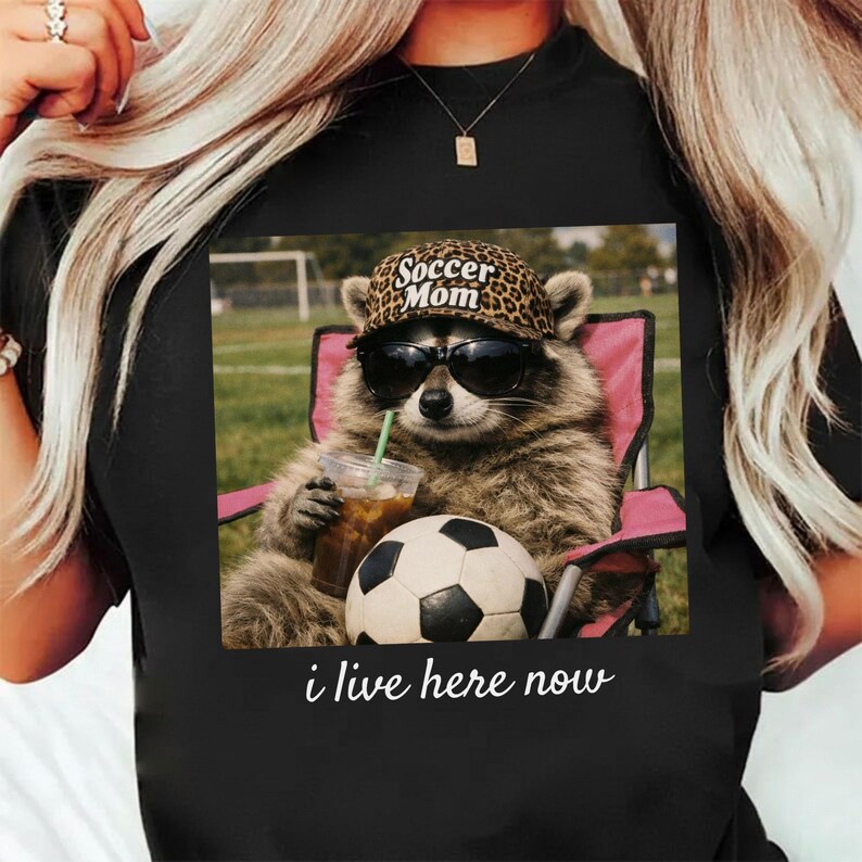 Puede incluir: Camiseta negra con un gr&aacute;fico de un mapache con gafas de sol y un sombrero de "Soccer Mom", sentado en una silla rosa, sosteniendo una bebida y un bal&oacute;n de f&uacute;tbol. El texto "i live here now" est&aacute; debajo.