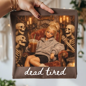 K&ouml;nnte beinhalten: Ein brauner Stoffartikel mit einem Grafikdruck einer blonden Puppe im grauen Kapuzenpullover, die in einem Samtsessel sitzt, einen Eiskaffee h&auml;lt und von zwei Skeletten flankiert wird. Der Text "dead tired" steht in wei&szlig;er Schrift unten.