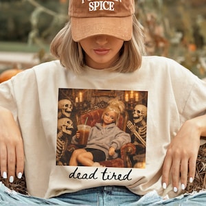 K&ouml;nnte beinhalten: Ein beiges T-Shirt mit einem Grafikdruck einer Puppe in einem grauen Kapuzenpullover, die in einem Sessel sitzt, einen Eiskaffee h&auml;lt und von zwei Skeletten umgeben ist. Der Text "dead tired" steht in schwarzer Schreibschrift darunter.