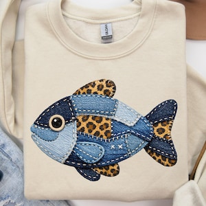 Puede incluir: Una sudadera color crema con un diseño de pez de mezclilla y estampado de leopardo. El pez está hecho de varios tonos de parches de mezclilla azul con acentos de estampado de leopardo. La sudadera tiene cuello redondo y mangas largas.