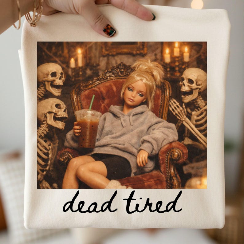 K&ouml;nnte beinhalten: Eine cremefarbene Tragetasche mit einem Grafikdruck einer blonden Puppe in einem grauen Kapuzenpullover und schwarzen Shorts, die in einem Samtsessel sitzt, einen Eiskaffee h&auml;lt und von zwei Skeletten flankiert wird, mit dem Text "dead tired".