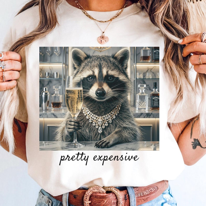 May include: Cream-colored t-shirt featuring a raccoon wearing a diamond necklace and holding a champagne flute. The raccoon is in a bar setting. The text "pretty expensive" is printed below the image.