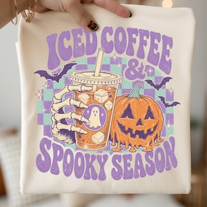 May include: A cream-colored t-shirt featuring a graphic print of a skeleton hand holding an iced coffee cup with a ghost icon, next to a melting orange pumpkin. The text reads ICED COFFEE & SPOOKY SEASON in purple, set against a teal and purple checkerboard.