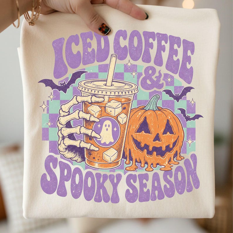 May include: A cream-colored t-shirt featuring a graphic print of a skeleton hand holding an iced coffee cup with a ghost icon, next to a melting orange pumpkin. The text reads ICED COFFEE & SPOOKY SEASON in purple, set against a teal and purple checkerboard.
