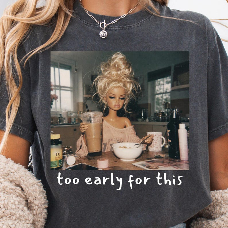 Peut inclure: Un t-shirt gris anthracite avec une impression graphique repr&eacute;sentant une poup&eacute;e aux cheveux blonds en chignon d&eacute;coiff&eacute;, assise &agrave; une table de cuisine avec un caf&eacute; et un bol de c&eacute;r&eacute;ales. Le texte en dessous indique "too early for this".