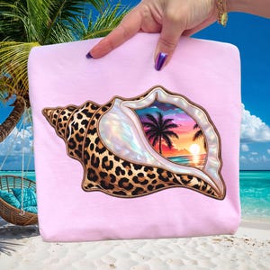 May include: A pale pink sweatshirt with a seashell design. The shell has a leopard print pattern and a pearlescent interior. Inside the shell is a tropical beach scene with palm trees, a sunset, and ocean waves.