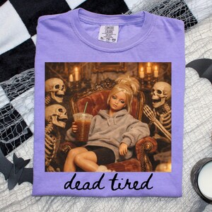 K&ouml;nnte beinhalten: Ein hellviolettes T-Shirt mit einem Grafikdruck einer blonden Puppe im grauen Kapuzenpullover, die in einem Sessel zwischen zwei Skeletten sitzt und einen Eiskaffee h&auml;lt. Der Text "dead tired" steht in schwarzer Schreibschrift darunter.