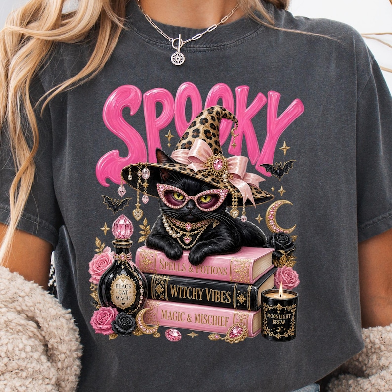 May include: A charcoal gray t-shirt featuring a graphic of a black cat wearing a leopard print witch hat and pink glasses. The cat rests on three books labeled Spells & Potions, Witchy Vibes, and Magic & Mischief, with a bottle and candle nearby.