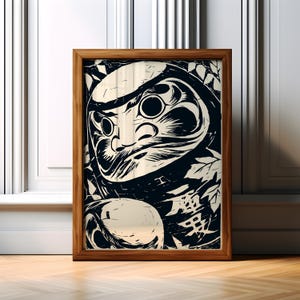 May include: A framed print featuring a Daruma doll illustration in black and cream. The artwork has a bold, graphic style with a focus on the doll's face and details. The wooden frame adds a classic touch to the piece.