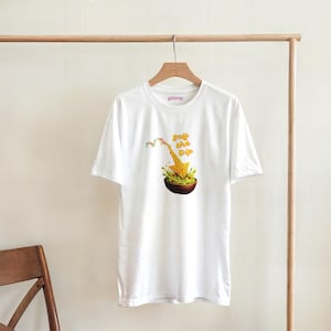 Pode incluir: Camiseta branca com um gráfico de um gráfico do mercado de ações e uma seta para baixo, uma tigela de guacamole e o texto "Buy the Dip" em amarelo. A camiseta está pendurada em um cabide de madeira.