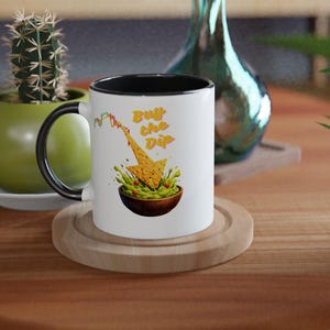 Pode incluir: Caneca de cerâmica branca com interior e alça pretos, com o texto "Buy the Dip" e um gráfico de um chip de tortilla mergulhando em guacamole. A caneca está sobre um descanso de madeira.