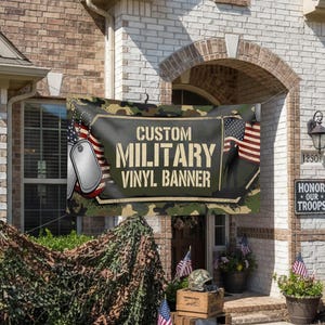 May include: A custom military vinyl banner is displayed above a doorway, with the text "Custom Military Vinyl Banner." The banner features dog tags and an American flag. The scene includes camouflage netting, wooden crates, and American flags.