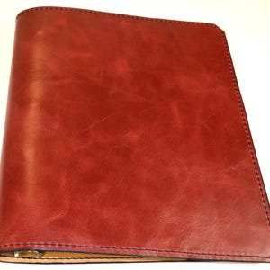 May include: A deep red binder with a smooth, slightly marbled texture and visible dark stitching along the edges. The cover features a rounded spine on the left side and a straight, angled cut at the bottom right corner, revealing a light tan interior.